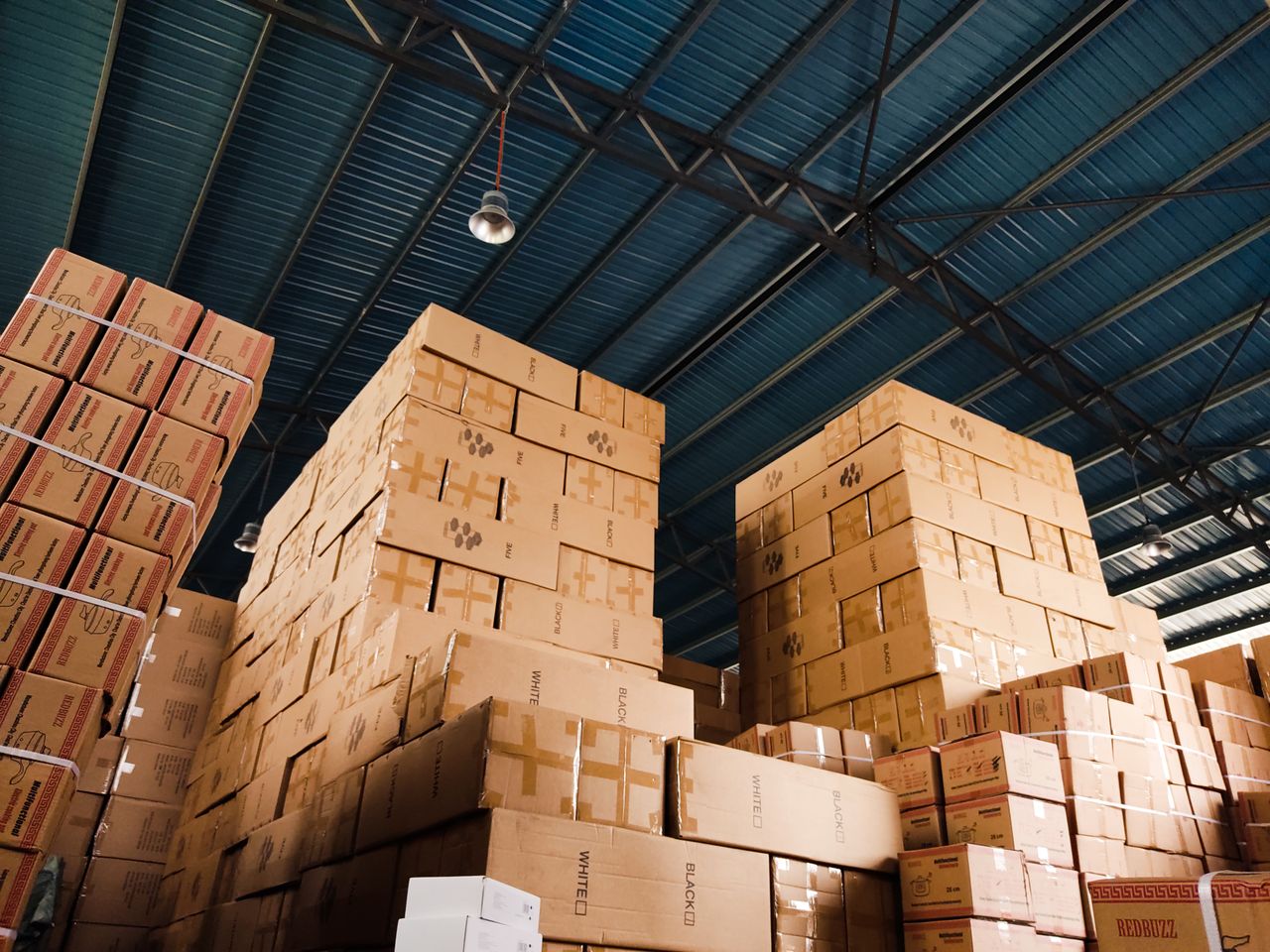 hero-img-01 High stacks of cardboard boxes organized in a warehouse with a blue metal ceiling.