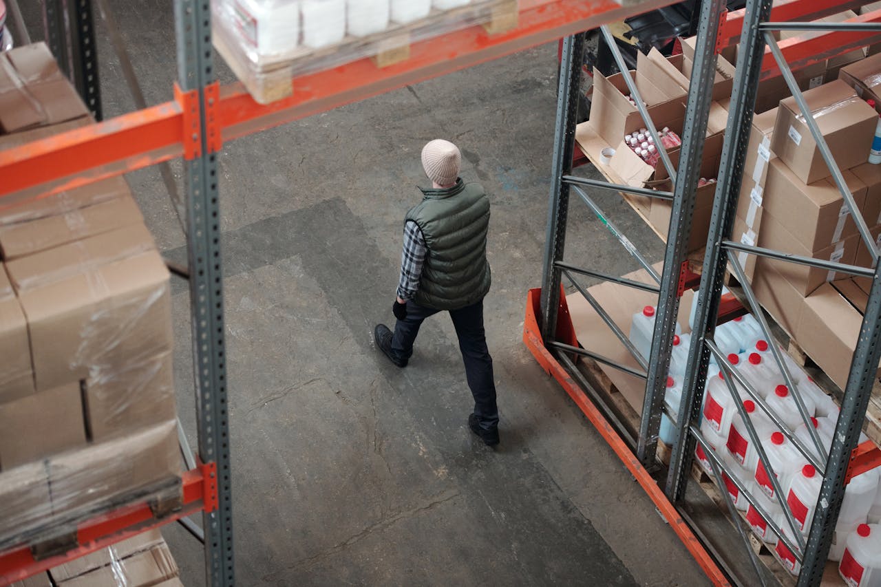 hero-img-02 Overhead view of a worker walking through a warehouse aisle surrounded by shelves of packages.