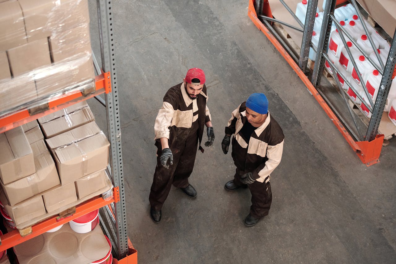 Two adult men in a warehouse communicating about storage logistics, surrounded by shelves of packages.